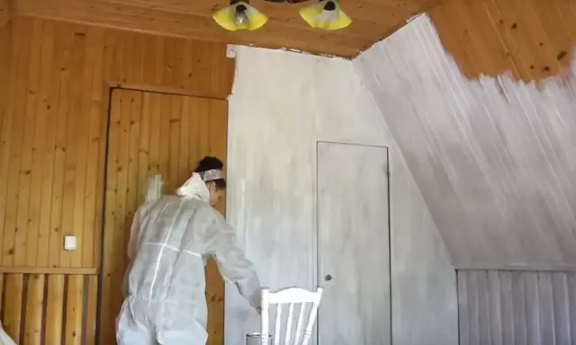 Osoba w kombinezonie maluje boazerię na biało. Prace remontowe w pokoju z drewnianym sufitem.
