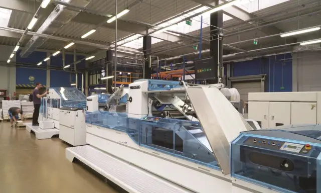 Wielka hala produkcyjna, gdzie pracuje najnowocześniejsza maszyna drukarska. Pracownicy nadzorują proces druku, który odbywa się w największej drukarni w Polsce.