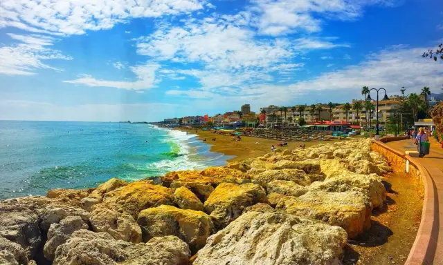 Sonniger Tag an der playa de la carihuela. Türkises Meer, goldener Sandstrand mit Liegen und Palmen, gesäumt von bunten Gebäuden. Felsen im Vordergrund.