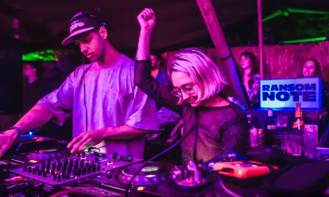 DJ set b2b: un uomo e una donna mixano musica in un locale con luci viola e verdi. La folla balla.