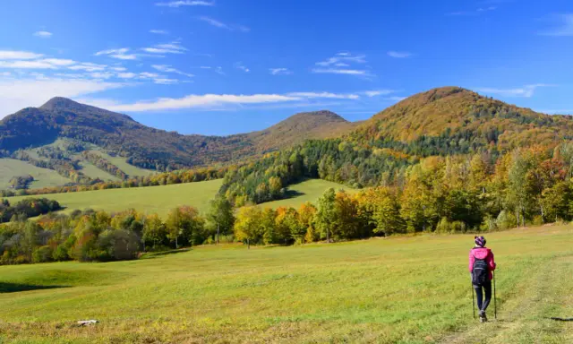 Jesień na **Beskid Niski szlaki**. Turystka w różowej kurtce idzie przez zieloną łąkę w kierunku kolorowych wzgórz.