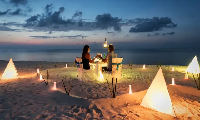 Una coppia al mare di sera, seduta a tavola per una cena romantica illuminata da lanterne e luci soffuse sulla sabbia.