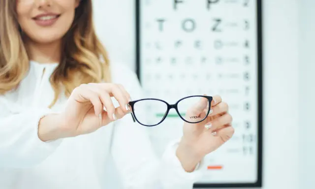 Okulary z astygmatyzmem – czy trzeba nosić cały czas? Optometrystka prezentuje okulary na tle tablicy Snellena.