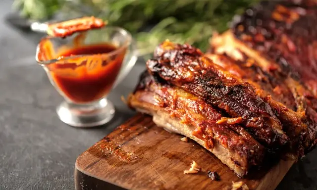Costine al forno succulente e caramellate, servite su tagliere di legno con salsa barbecue in ciotolina.
