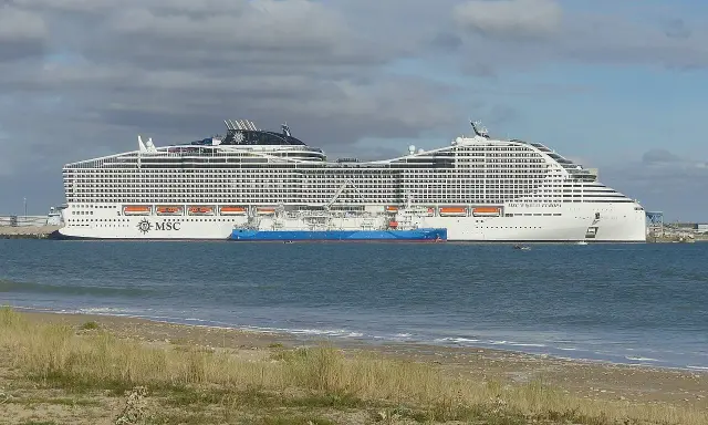 A large white MSC World Europa cruise ship docked next to a blue tanker ship in the sea.