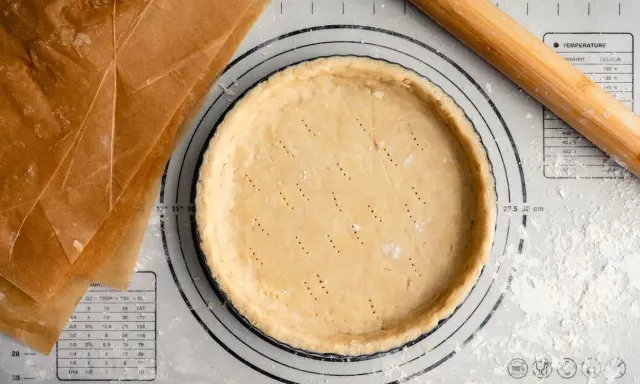 Pâte sucrée noisette prête à être garnie, sur un tapis de pâtisserie avec un rouleau à bois et du papier sulfurisé.