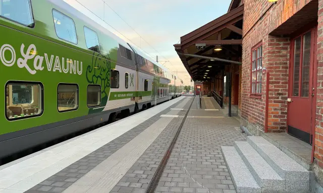 Grönt tåg med texten "LaVaunu" står vid en tegelbyggnad. Kanske är det ett tåg till Haparanda som väntar på passagerare.