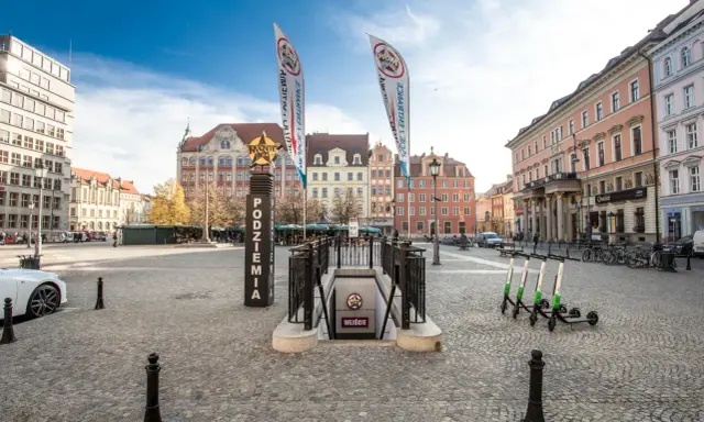 Wejście do podziemi z napisem "Movie Gate" i dwoma pionowymi banerami. W tle kolorowe kamienice i błękitne niebo.