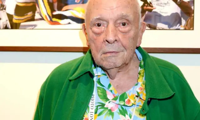 An elderly man with a green jacket over a floral shirt. This image might spark some david bailey interesting facts about his iconic style.