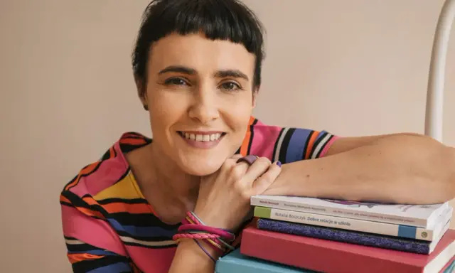 Uśmiechnięta kobieta z krótkimi włosami opiera się o stos kolorowych książek. Wiedza, którą posiadają, jest dla niej dostateczna.
