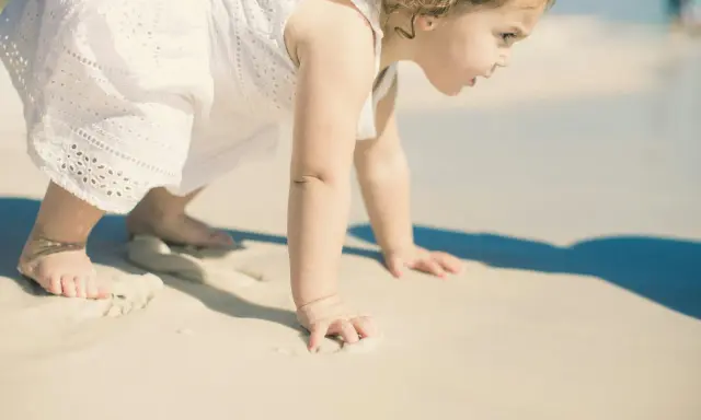 Małe dziecko w białej sukience raczkuje po plaży. Może to być jedna z przyczyn nie chodzenia dziecka, bo piasek jest ciekawy.