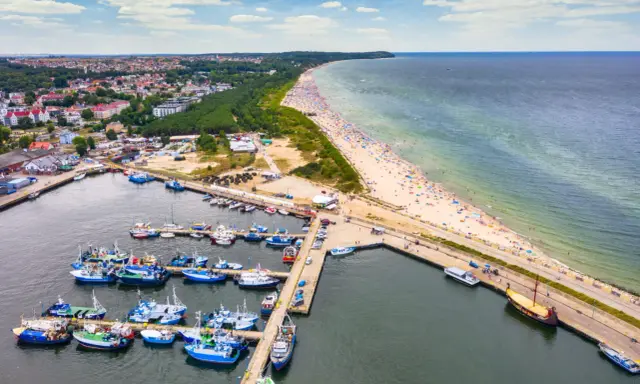 Władysławowo co zobaczyć: port z łodziami, szeroka plaża pełna turystów i miasto w tle. Idealne miejsce na wakacje nad morzem.