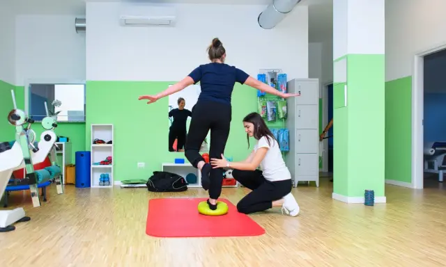 Kobieta ćwiczy propriocepcję na poduszce balansującej, druga osoba asekuruje.