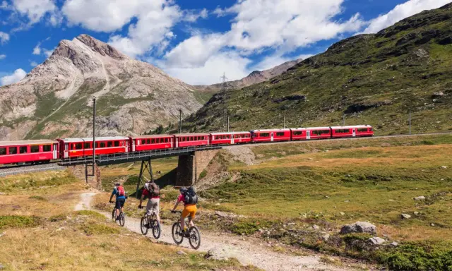 Treno rosso attraversa un ponte su un paesaggio alpino mozzafiato. Tre ciclisti pedalano su un sentiero sottostante, godendosi la vista della ferrovia del Bernina.