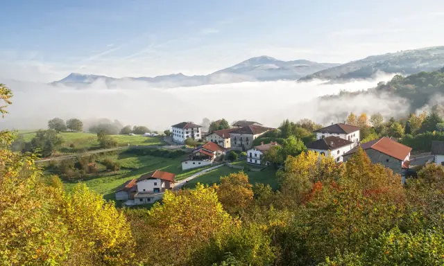 Pueblo en el **valle del Baztan** cubierto por niebla matutina, con casas blancas y tejados rojos entre árboles otoñales.