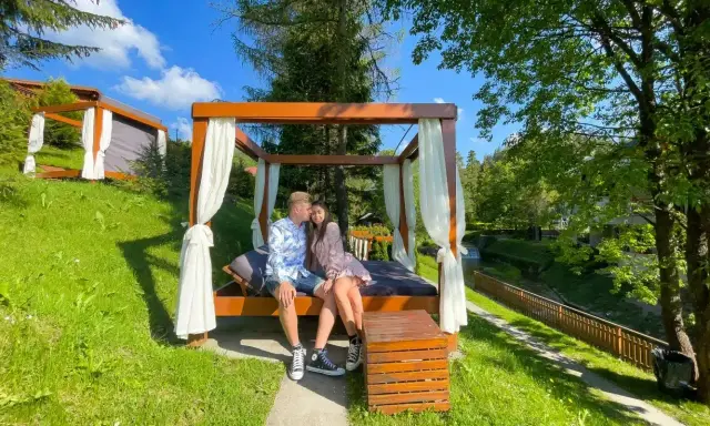 Para zakochanych cieszy się chwilą podczas wyjazdu na weekend. Siedzą na łóżku pod baldachimem, otoczeni zielenią i strumykiem.