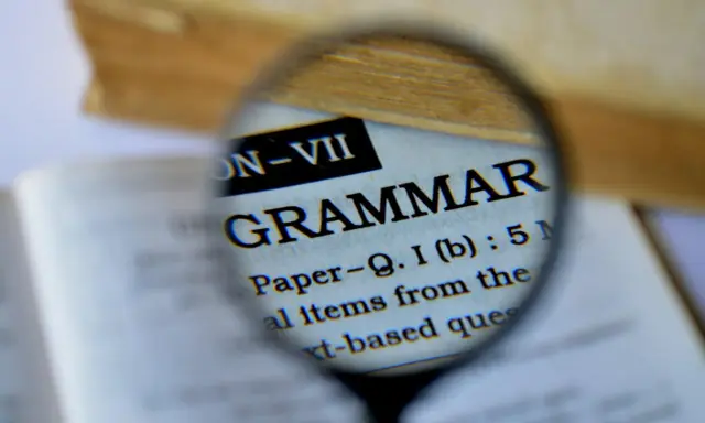 Une loupe examine le mot "GRAMMAR" dans un livre. Pour maîtriser la langue, il faut bien receler la conjugaison.