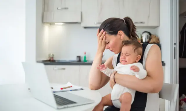 Zmęczona mama z płaczącym niemowlakiem na rękach, zastanawia się, przebodźcowanie niemowlaka ile trwa. Obok laptop i dokumenty.