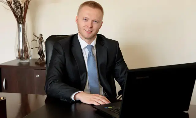 Mężczyzna w garniturze siedzi przy biurku z laptopem, obok statuetka Temidy.