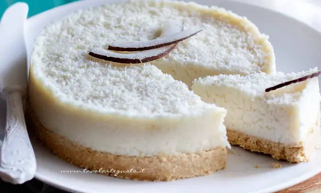 Delizioso cheesecake al cocco, ricoperto di cocco grattugiato e decorato con scaglie di cocco fresco. Una fetta è già stata tagliata.