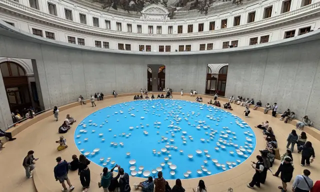 A circular room with a frescoed ceiling, featuring a bright blue pool dotted with white ceramic bowls. Visitors observe this installation, part of the François-Henri Pinault art collection.