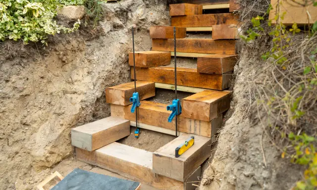 Construction d'un escalier en bastaing dans une pente. Des serre-joints bleus maintiennent les poutres en bois pendant que le niveau à bulle assure la précision.
