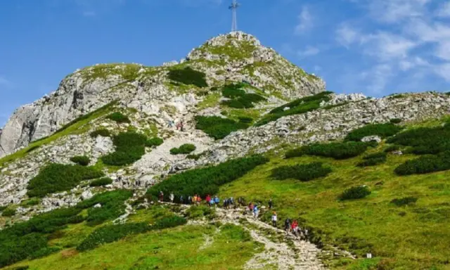Grupa turystów wchodzi kamienistym szlakiem na zielone zbocze góry z krzyżem na szczycie.