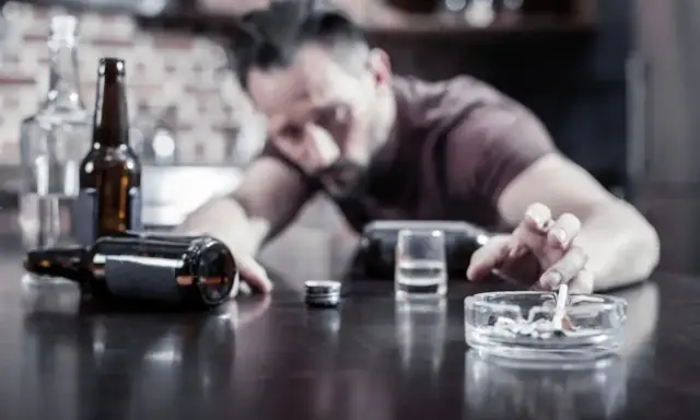 Mężczyzna z papierosem w popielniczce, butelki i kieliszek na stole. Nadmierne spożycie alkoholu może pogorszyć hemoroidy.