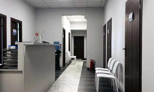 Korytarz kliniki medycznej z recepcją, drzwiami i białymi krzesłami.