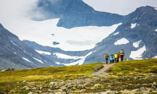 Vandringsgrupp på väg uppför en gräsbeklädd sluttning med stenar, mot en glaciärklädd bergstopp. En spännande utmaning att bestiga Sylarna.