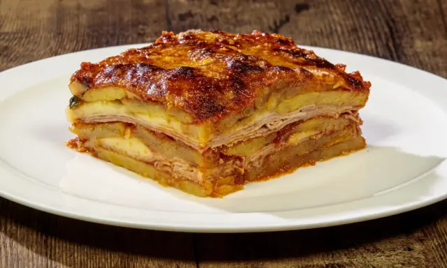 Una fetta di parmigiana di patate, stratificata con formaggio e salumi, servita su un piatto bianco.