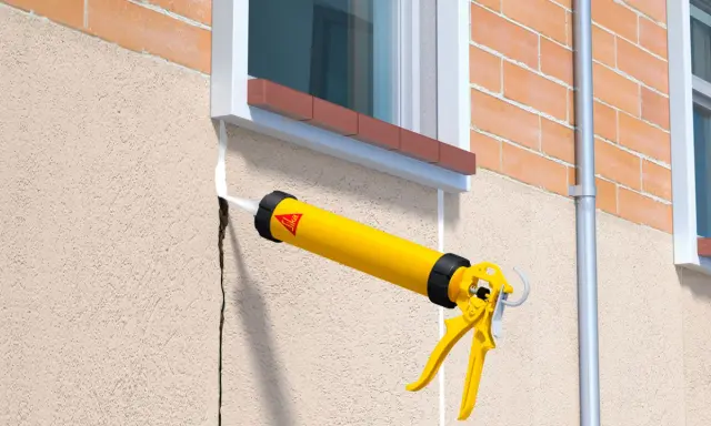 Pistola de calafateo amarilla aplicando sellador blanco para como tapar grietas en la pared exterior de estuco junto a una ventana.