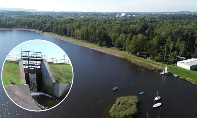 Widok na zbiornik wodny z zaporą i spływającą wodą. Na wodzie kilka łodzi, w tle las i zabudowania. To piękne **chechło** jest idealnym miejscem do wypoczynku.