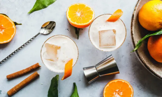 Zwei cremige Gin Sours mit Eis und Orangenzesten, garniert mit Zimtstangen und frischen Mandarinen.