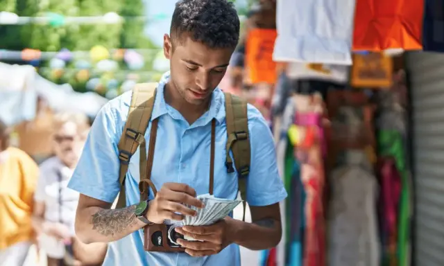 Mężczyzna z plecakiem liczy banknoty dolarów na tle kolorowego straganu.