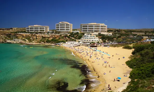 En livlig strand med turkost vatten och gyllene sand vid Golden Bay, Malta. Flera hotell syns i bakgrunden.