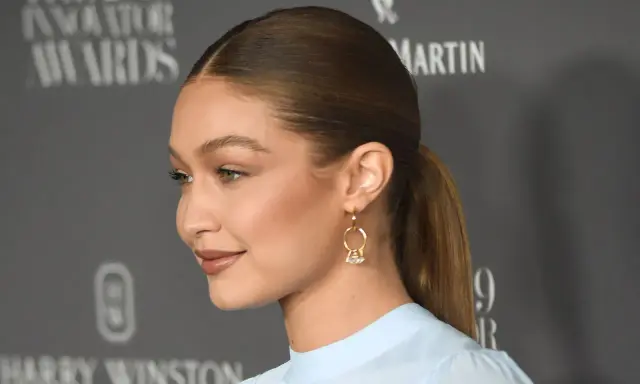 Gigi Hadid mit glatten, **haare nach hinten** gebundenen Haaren und einem dezenten Lächeln. Sie trägt ein hellblaues Oberteil und auffällige Ohrringe.