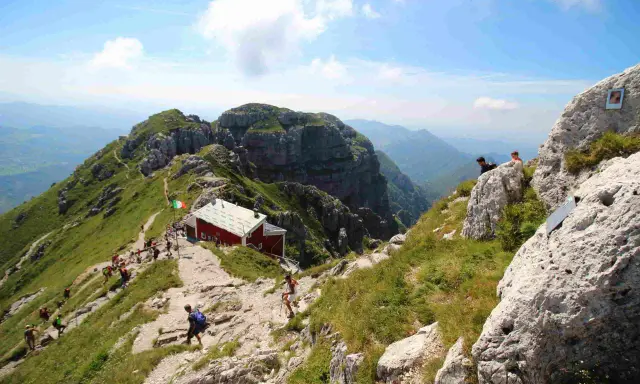 Sentiero panoramico verso il rifugio Lecco, con escursionisti su un crinale erboso e roccioso sotto un cielo azzurro.