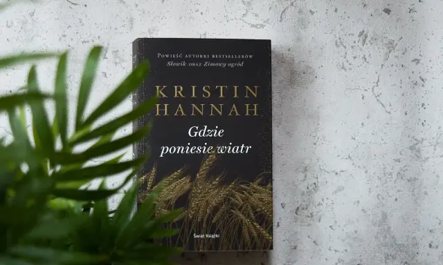 Okładka książki Kristin Hannah "Gdzie poniesie wiatr" na tle szarej ściany, z zielonymi liśćmi rośliny w zbliżeniu.