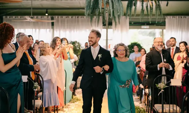 El novio sonriente camina del brazo de su madrina, ambos sonríen a los invitados.