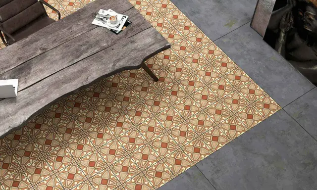 Un suelo hidráulico con diseño geométrico en tonos tierra y ocre, junto a un suelo de cemento pulido. Una mesa de madera rústica y una silla completan la escena.