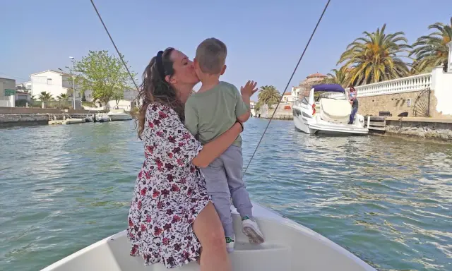 Madre besa a su hijo en un paseo en barco por los canales de Empuriabrava, con palmeras y casas blancas al fondo.