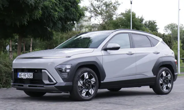 Nowy Hyundai Kona Hybrid w kolorze jasnoszarym, zaparkowany na brukowanej nawierzchni, otoczony zielenią.