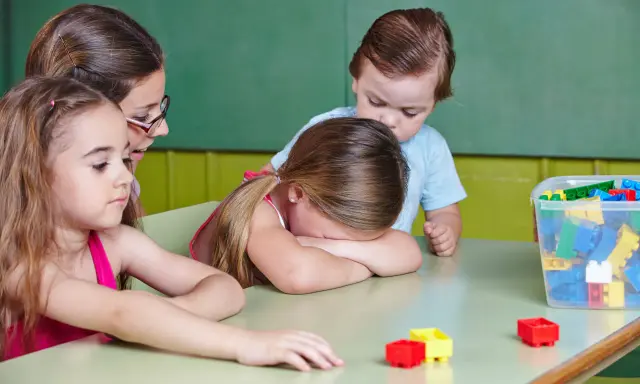 Smutna dziewczynka z płaczem ukrywa twarz na stole, podczas gdy inne dzieci i nauczycielka próbują ją pocieszyć. To obraz nieudanej adaptacji w przedszkolu.