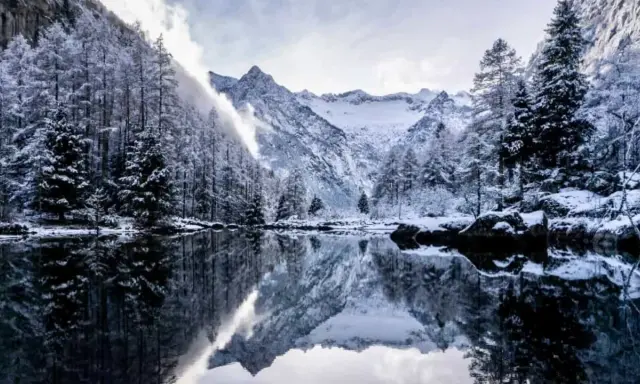 Val di Mello inverno: alberi innevati e montagne maestose si specchiano in un lago calmo, creando un paesaggio da fiaba.