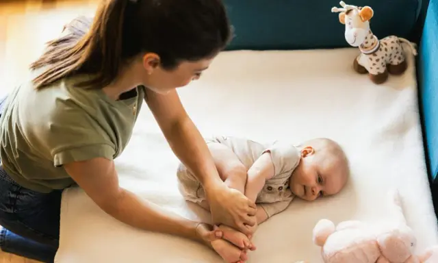 Mama bawi się z 3 miesięcznym dzieckiem, delikatnie masując jego stópki. Obok leżą pluszowe zabawki.