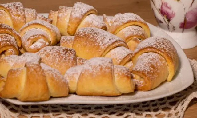 Talerz pełen puszystych, posypanych cukrem pudrem rogalików krucho drożdżowych. Idealne na śniadanie lub deser.