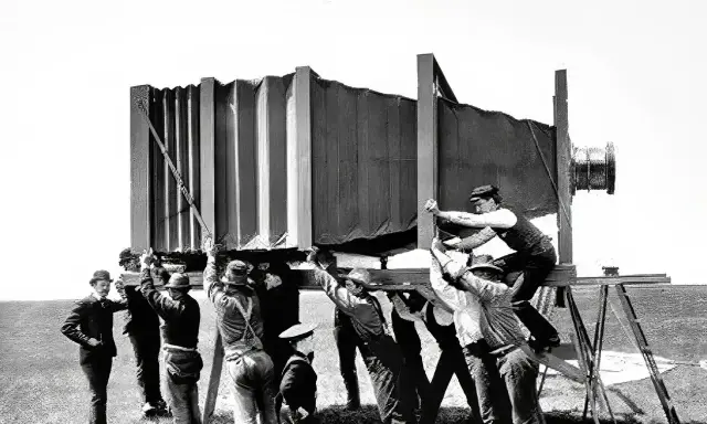 Grupa mężczyzn w historycznych strojach ustawia gigantyczny aparat fotograficzny. Wyobraź sobie, kiedy wynaleziono aparat fotograficzny i jak wielkie musiały być pierwsze urządzenia!