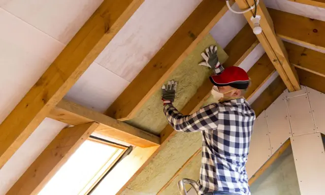 Uomo con mascherina e guanti installa materiale isolante tra le travi di legno di un sottotetto. È chiaro che conviene isolare il sottotetto per migliorare il comfort.