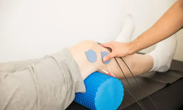 Rehabilitacja kolana w domu: elektrody na skórze, wałek do masażu pod nogą, ręka terapeuty.